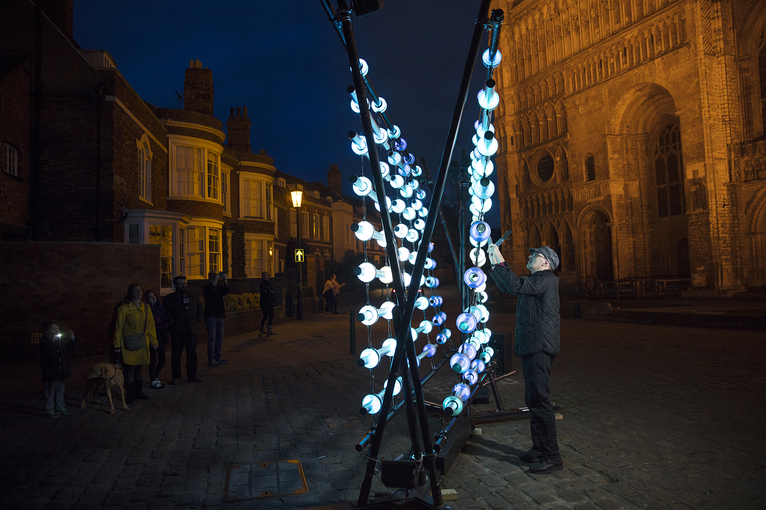 A man plays the Illumaphonium outside a church. It is a large sculpture made of glass lights and pipes which he is hitting with special sticks.