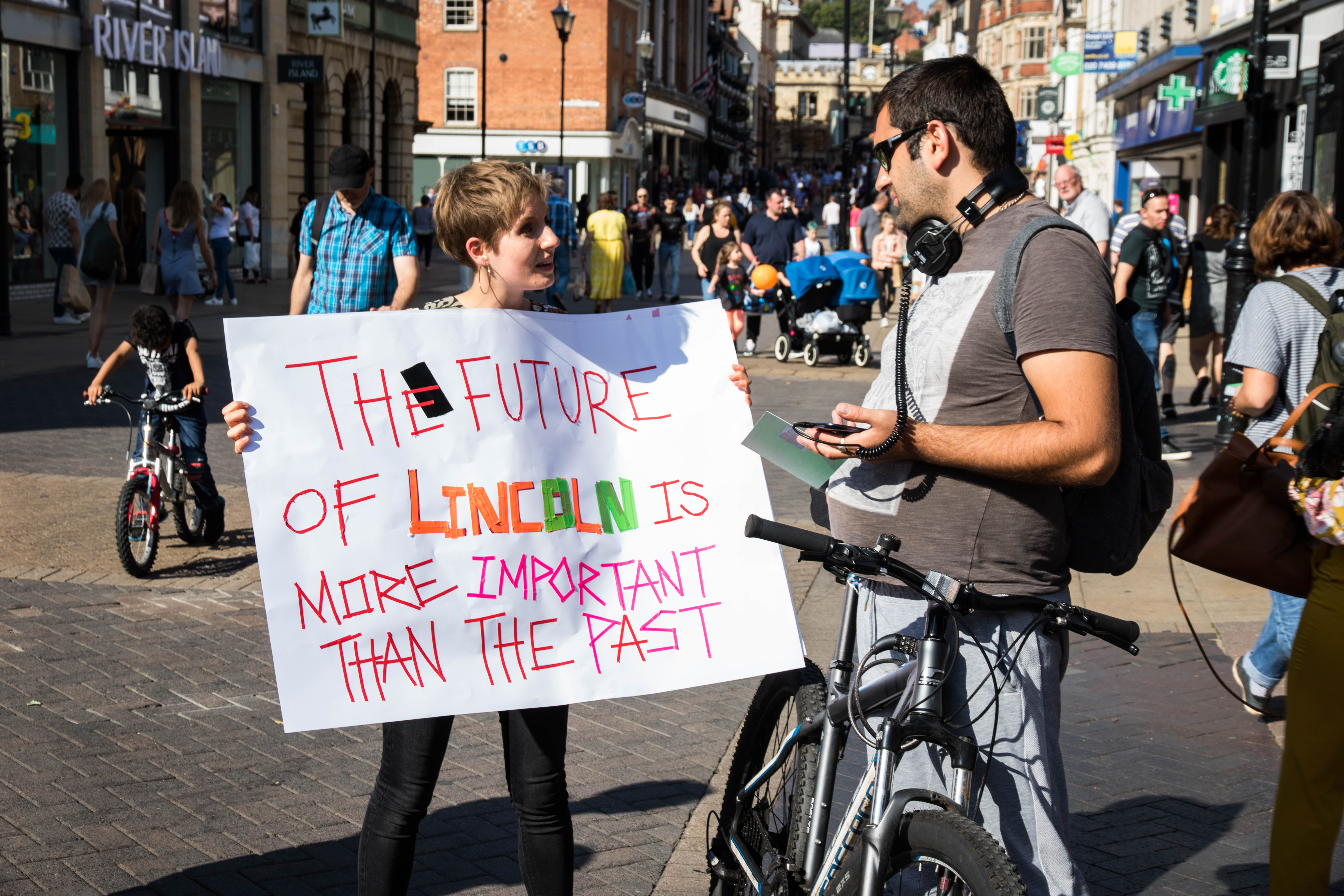 In the street a young woman holds a sign up as she talks to a man with a bicycle. It reads The future of lincoln is more important than the past.