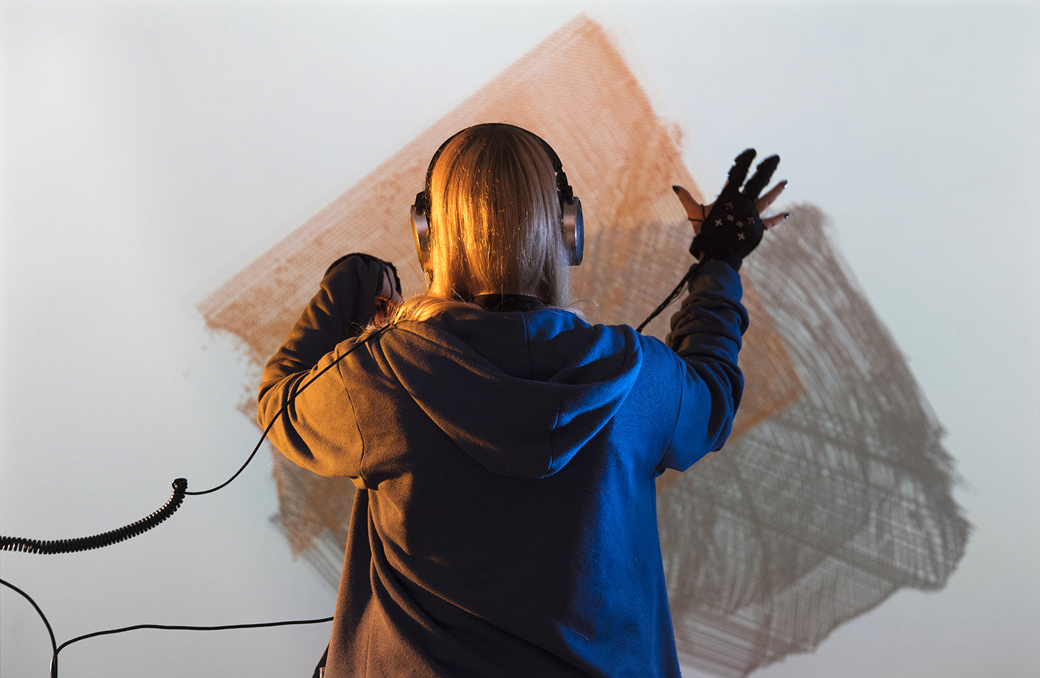 A woman is seen from behind, she wears a wired headset and gloves and is drawing with her hards on a digital screen