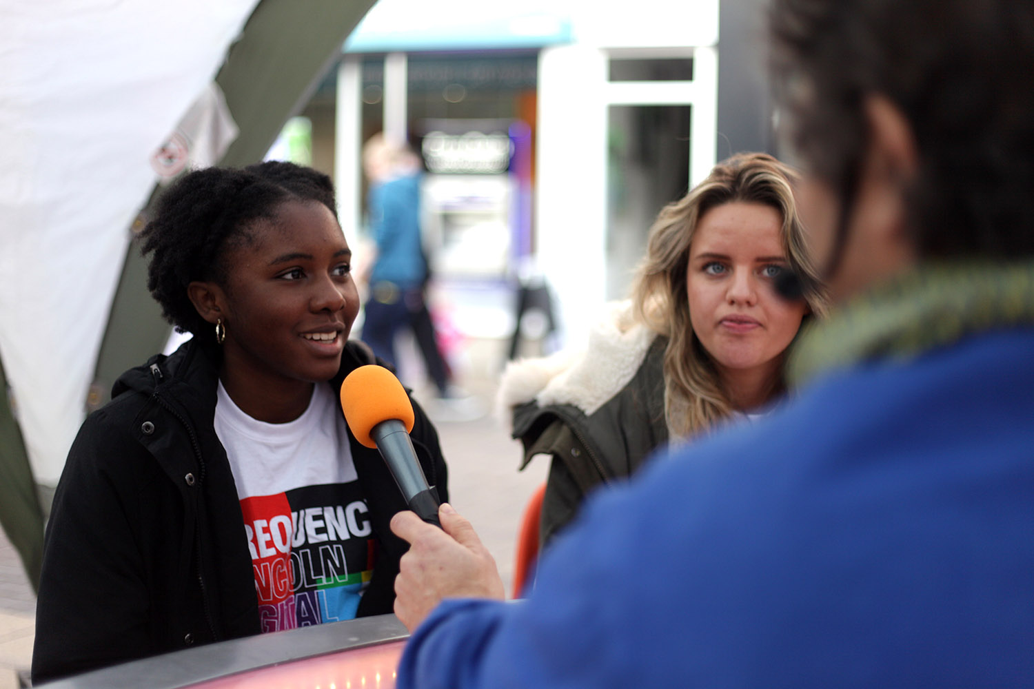 Two young women, one black one white, are speaking into a microphone for an interview in the street.