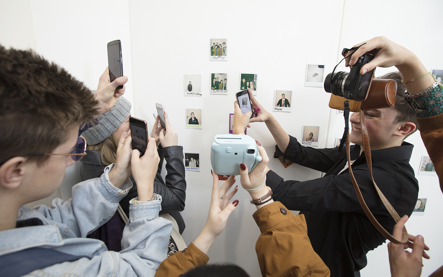 A group of young people hold up different types of cameras to an exhibit of polaroid photographs