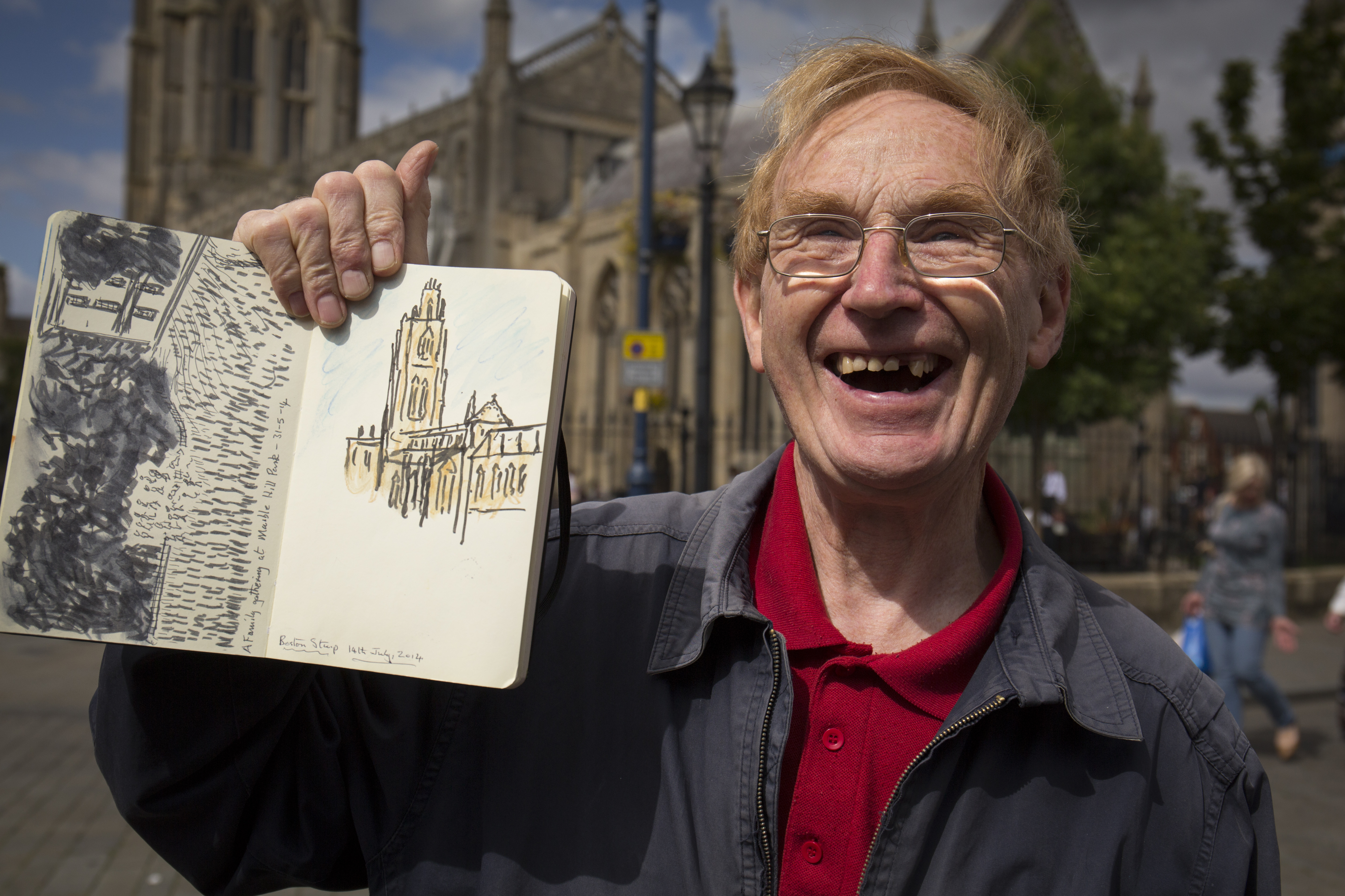 An older man holds up a drawing he has made of Lincoln Cathedral and smiles joyfully