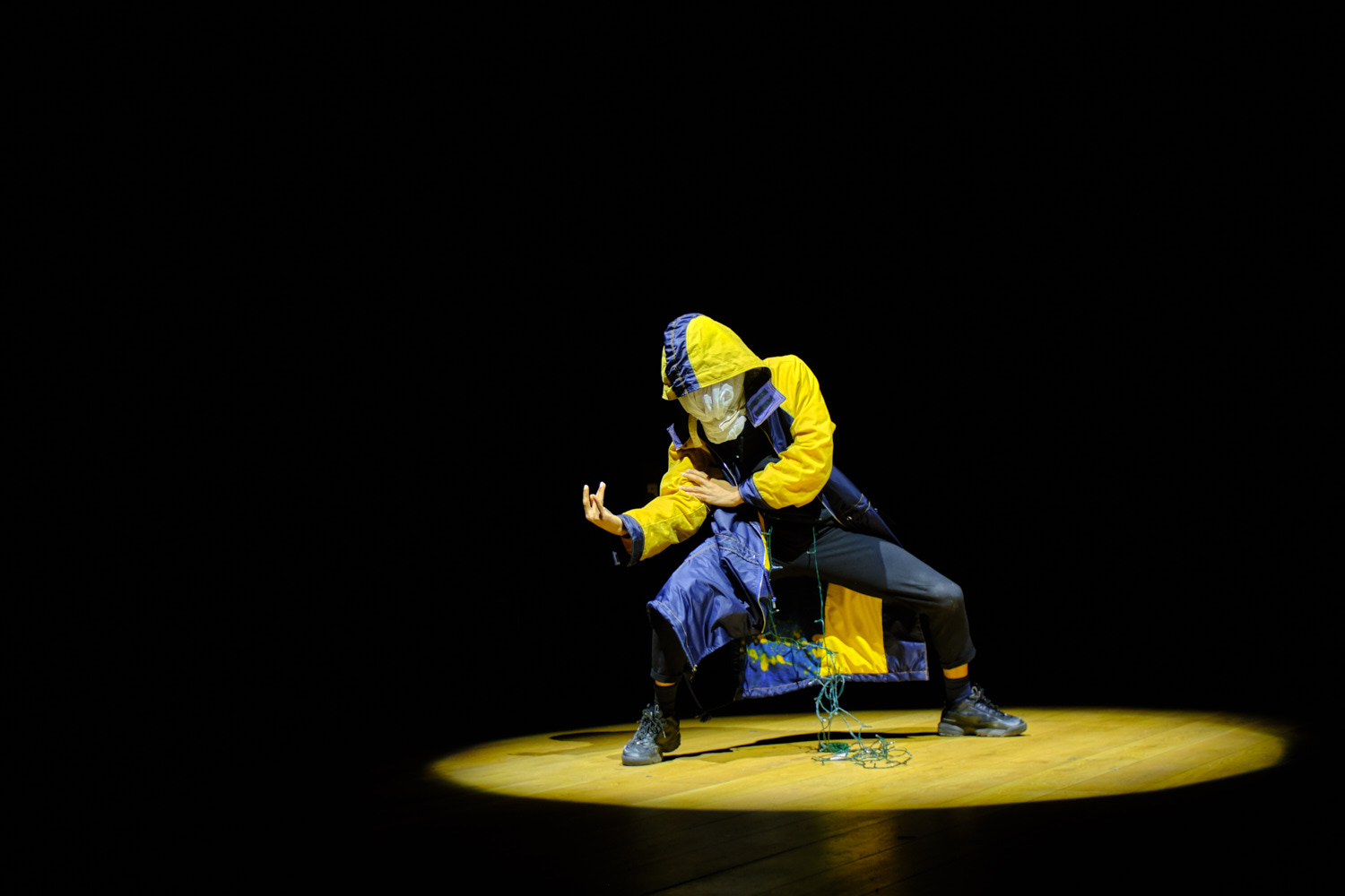 A young artist from in the UK dances, spotlit on a black stage wearing a mask and a blue and yellow coat.