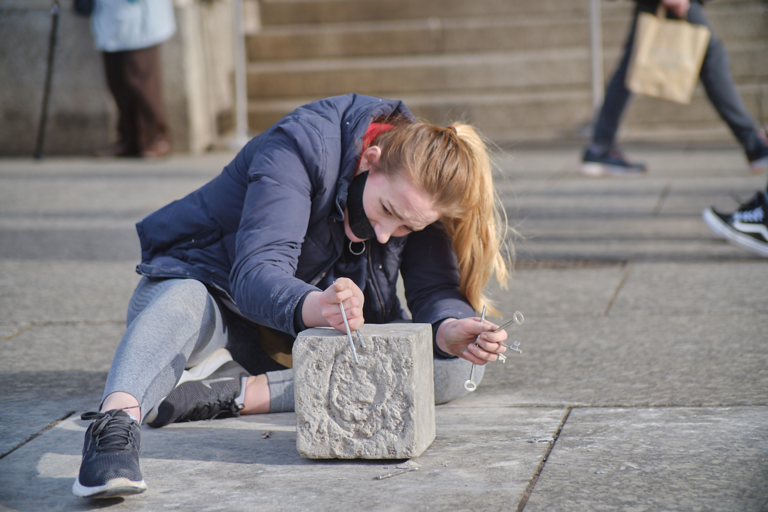 An artist performs with black tape on her mouth, she sits on the floor and carves a block of stone with keys.