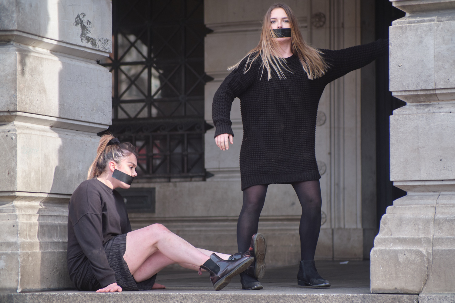 Two female performers are in a public space with black tape covering their mouths, wearing black outfits.