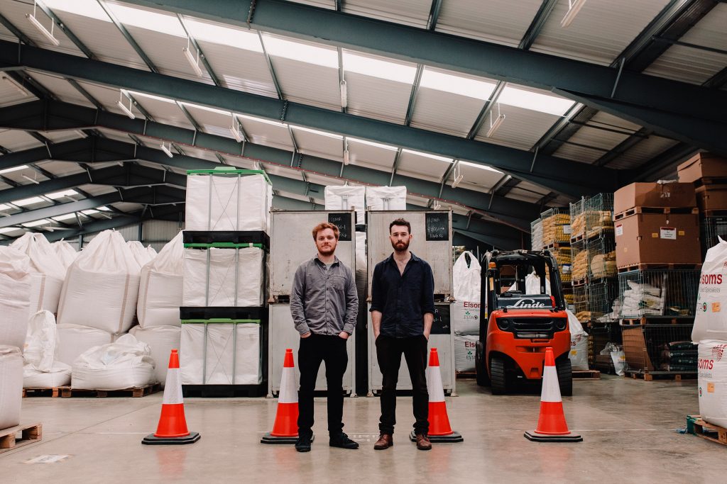 Two young white men stand with hands in pockets in a warehouse, surrounded by cones and industrial machines