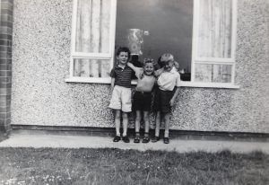 An old photograph where three young boys stand with their arms around each other, in short trousers outside a pebble-dashed house.